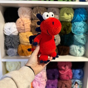 Puede incluir: Un juguete de dinosaurio de crochet rojo con ojos azules y garras negras. El juguete está siendo sostenido en una mano frente a un estante con ovillos de lana de colores.