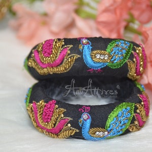 May include: Two black fabric bangles with peacock and floral embroidery. The bangles are decorated with gold thread, pink, blue, and green embroidery. The bangles are stacked on top of each other.