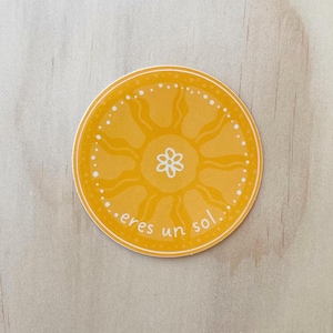 May include: A round, yellow sticker with a sun design and the text "eres un sol." The sun has wavy rays and a white flower in the center. White dots encircle the sun's edge. The sticker is on a wooden surface.
