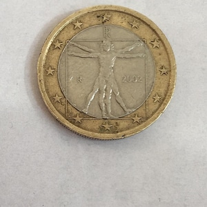 Puede incluir: Una moneda de un euro con el Hombre de Vitruvio, un dibujo de Leonardo da Vinci, fechado en 2002. La moneda tiene un anillo exterior dorado con estrellas y un centro plateado con la ilustración.