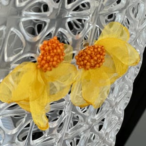 May include: A pair of yellow flower earrings with orange beaded centres. The earrings are set against a clear, textured background with a geometric pattern. The flowers have delicate, translucent petals.