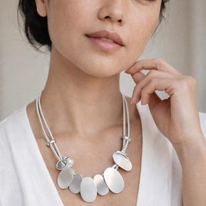 May include: A silver necklace featuring oval and circular pendants. The necklace has a double-strand design and is worn against a white top. The pendants are matte silver and arranged in a cascading pattern.