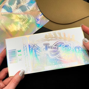 May include: A white ticket with a holographic finish. The ticket says "Theme Park" and has a barcode that says "KIE" and a number. The ticket also says "Day Pass" and "Official Date & Time".