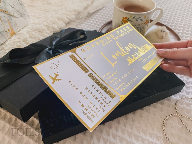 May include: A gold boarding pass with the text "Boarding Pass Happy Mother's Day London" and "Passenger: MUMMA Departure: 11TH AUG 2022 From: ABERDEEN Duration: 2 NIGHTS Booking Ref: MOTHERSDAY 1176 186 187" The boarding pass is in a black box with a black ribbon.