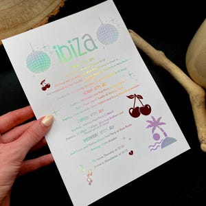 May include: A white paper with the word "ibiza" in iridescent lettering, featuring a schedule of events. The design includes disco balls, cherries, a palm tree, and musical notes. The paper is held by a hand.