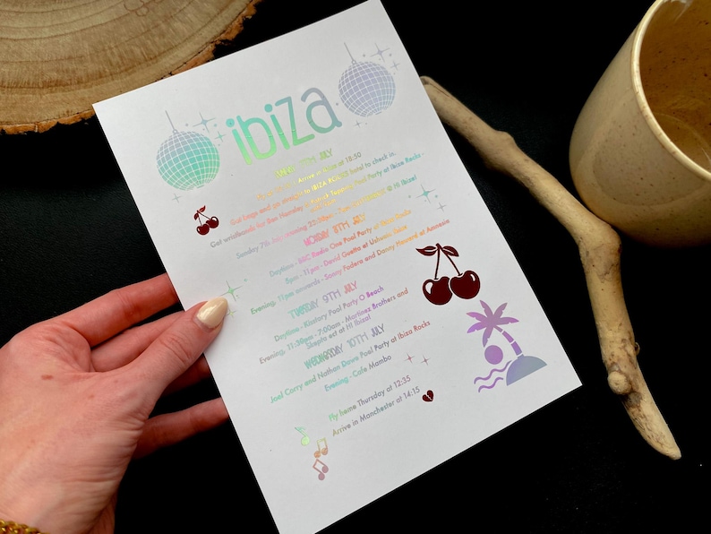 May include: A white paper with the word "ibiza" in iridescent lettering, featuring a schedule of events. The design includes disco balls, cherries, a palm tree, and musical notes. The paper is held by a hand.