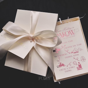 May include: Ivory-colored gift box tied with a satin ribbon bow. A card with gold lettering that says "Happy Birthday" is attached. An open card displays a pink and white travel itinerary with the text "Winter Holiday in RAKOW".