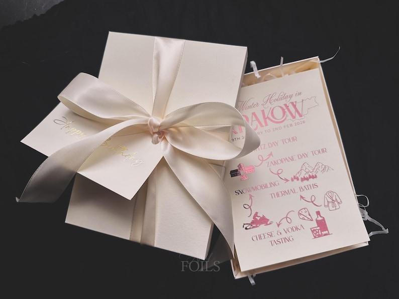 May include: Ivory-colored gift box tied with a satin ribbon bow. A card with gold lettering that says "Happy Birthday" is attached. An open card displays a pink and white travel itinerary with the text "Winter Holiday in RAKOW".