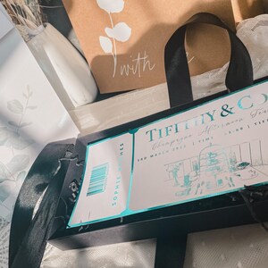 May include: A black box with a Tiffany & Co. Champagne Afternoon Tea invitation card. The card is light blue and white with a drawing of a tea room. The text on the card reads "Tiffany & Co. Champagne Afternoon Tea 3rd March 2023 | Time 2:00 | Tiffany's"