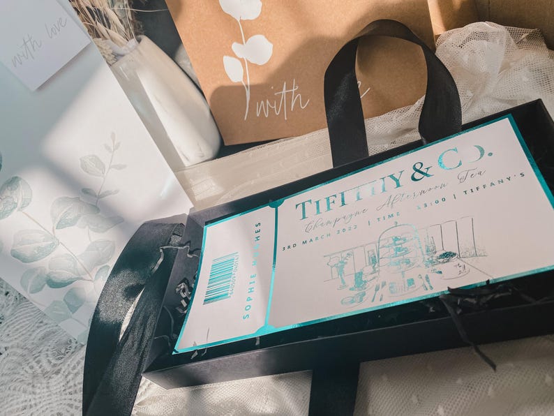 May include: A black box with a Tiffany & Co. Champagne Afternoon Tea invitation card. The card is light blue and white with a drawing of a tea room. The text on the card reads "Tiffany & Co. Champagne Afternoon Tea 3rd March 2023 | Time 2:00 | Tiffany's"