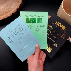 May include: Three travel itinerary cards are displayed. One is light blue with "ICELAND" and a compass rose. A green card says "BARCELONA" with illustrations. A black card says "ROME" with a date. The cards are held by a hand.