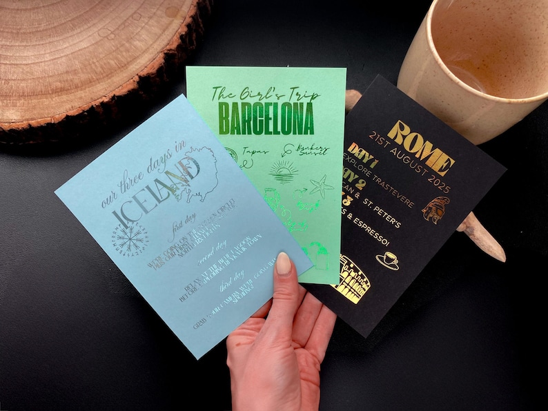 May include: Three travel itinerary cards are displayed. One is light blue with "ICELAND" and a compass rose. A green card says "BARCELONA" with illustrations. A black card says "ROME" with a date. The cards are held by a hand.