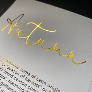 May include: A close-up of a white card with the word "Autumn" in gold script. The card also includes text describing the origin and meaning of the name "Autumn".