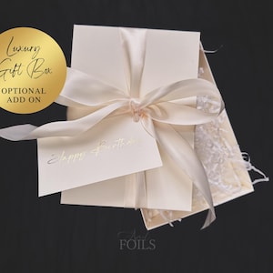 May include: A cream-colored gift box tied with a satin ribbon, with a small card that reads "Happy Birthday" in gold lettering. A gold circle with the words "Luxury Gift Box" and "Optional Add On" is in the upper left corner.