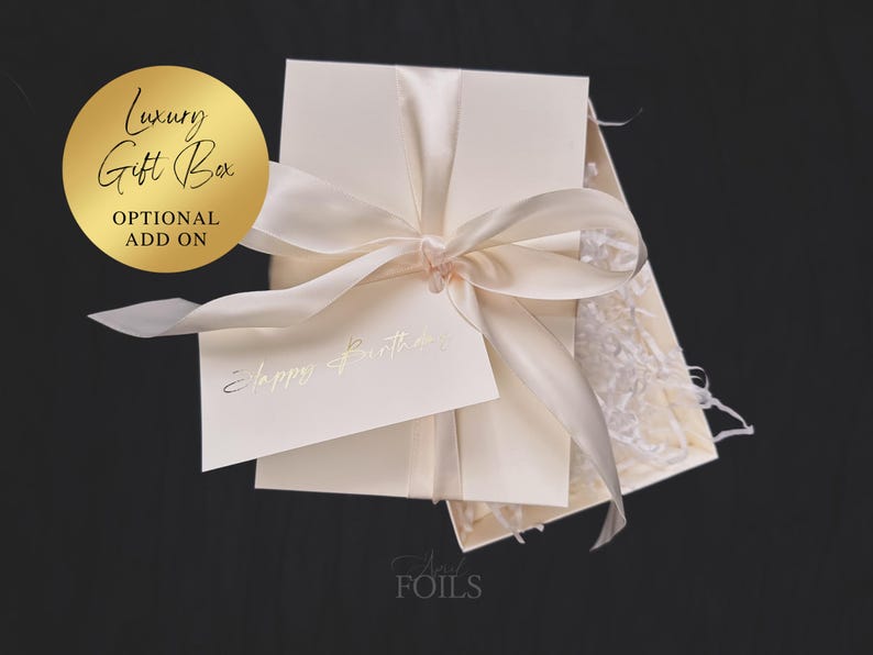 May include: A cream-colored gift box tied with a satin ribbon, with a small card that reads "Happy Birthday" in gold lettering. A gold circle with the words "Luxury Gift Box" and "Optional Add On" is in the upper left corner.