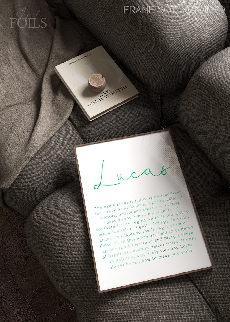 May include: A framed print with the name "Lucas" in a stylish green script. The print includes a description of the name's origin and meaning. A book titled "at 100 A Century of Style" and a decorative object rest on a gray sofa.
