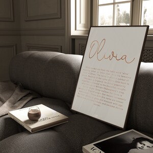 May include: A framed print with the name "Olivia" in copper-colored script. The print includes a description of the name's origin and meaning. The print is displayed on a gray sofa with books and a glass of liquid.