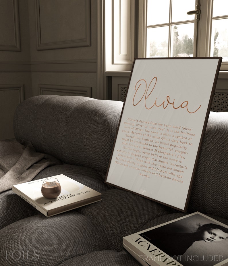 May include: A framed print with the name "Olivia" in copper-colored script. The print includes a description of the name's origin and meaning. The print is displayed on a gray sofa with books and a glass of liquid.