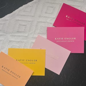 May include: A collection of five rectangular business cards in shades of peach, yellow, light pink, medium pink, and dark pink. Each card features the printed text KATIE ENGLER and RESTAURANT GROWER in a dark, serif font centred on the surface.