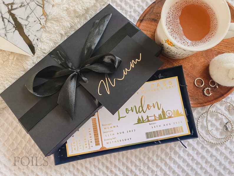 May include: A black gift box with a gold ribbon and a gold tag that says "Mum". The box is open and inside is a white card with a gold border and the text "London Pass - Happy Mother's Day!" with a gold illustration of the London skyline.