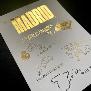 May include: A white card with gold foil text and illustrations. The word "MADRID" is at the top. Other text includes "Real Madrid vs Villarreal Santiago Bernabeu", "Hospitality", "Amazing Pastries!", and "Lots of pasta & wine at Circolo Popolare".