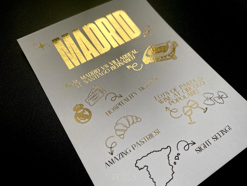May include: A white card with gold foil text and illustrations. The word "MADRID" is at the top. Other text includes "Real Madrid vs Villarreal Santiago Bernabeu", "Hospitality", "Amazing Pastries!", and "Lots of pasta & wine at Circolo Popolare".