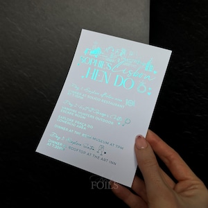 May include: A white invitation card with teal foil lettering. The card reads "Sophie's Lisbon Hen Do" and lists event details. The card is held by a person's hand against a dark background.