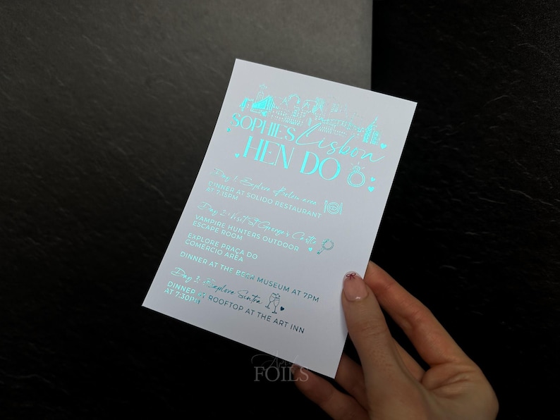 May include: A white invitation card with teal foil lettering. The card reads "Sophie's Lisbon Hen Do" and lists event details. The card is held by a person's hand against a dark background.