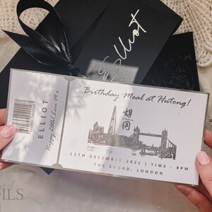 May include: A silver ticket with black text that reads "ELLIOT Happy 26th! Love M*" and "Birthday Meal at Hutong!" with a black and white illustration of the Shard and Tower Bridge in London. The text also reads "12TH DECEMBER 2021 | TIME - 8PM THE SHARD, LONDON".