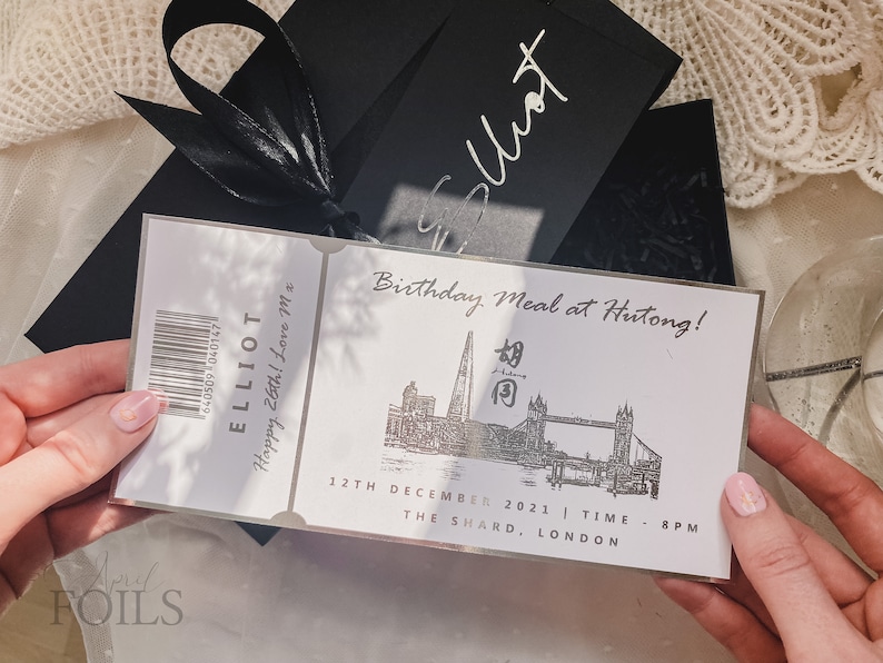 May include: A silver ticket with black text that reads "ELLIOT Happy 26th! Love M*" and "Birthday Meal at Hutong!" with a black and white illustration of the Shard and Tower Bridge in London. The text also reads "12TH DECEMBER 2021 | TIME - 8PM THE SHARD, LONDON".