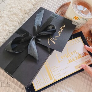 May include: A black gift box with a black ribbon and a gold inscription that says "Mum". The box is sitting on a white fuzzy surface. A white card with gold lettering says "London Pass Mother's Day!" and has a gold image of the London Eye.