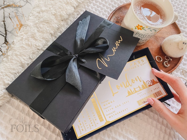 May include: A black gift box with a black ribbon and a gold inscription that says "Mum". The box is sitting on a white fuzzy surface. A white card with gold lettering says "London Pass Mother's Day!" and has a gold image of the London Eye.