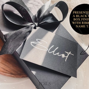 May include: A black gift box with a black satin ribbon and a black card with the word "Elliot" written in white. The box is presented in a black card box finished with ribbon and name tag.