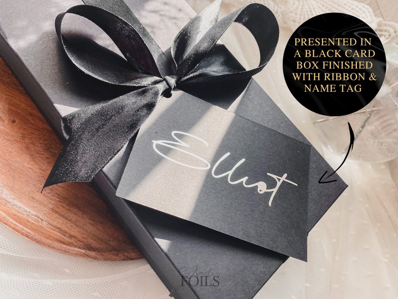 May include: A black gift box with a black satin ribbon and a black card with the word "Elliot" written in white. The box is presented in a black card box finished with ribbon and name tag.