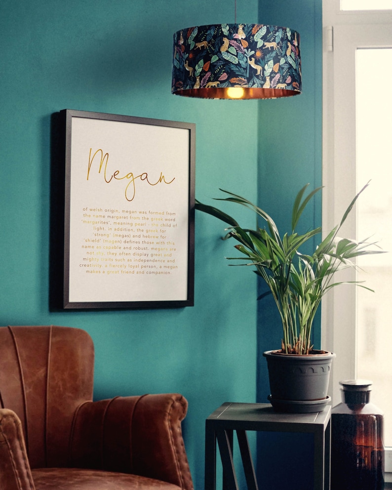 May include: A framed print with the name "Megan" in gold script, accompanied by a definition. Above, a lampshade with a dark blue background and a pattern of leaves and animals hangs. A brown leather armchair and a potted plant are also visible.