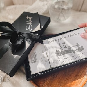 May include: A black gift box with a black ribbon tied around it. The box is open and inside is a white card with the text "Birthday Meal at Hutong!" and a black and white illustration of the London skyline. The card also includes the date "12th December 2021" and the time "8PM". The card is in a black box with a clear plastic cover.