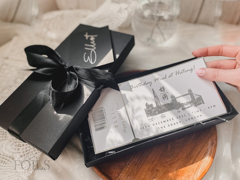 May include: A black gift box with a black ribbon tied around it. The box is open and inside is a white card with the text "Birthday Meal at Hutong!" and a black and white illustration of the London skyline. The card also includes the date "12th December 2021" and the time "8PM". The card is in a black box with a clear plastic cover.