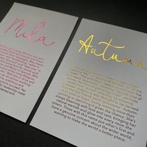 May include: Two cards with names printed in metallic foil. One card says "Mila" in pink, the other says "Autumn" in gold. Both cards have text describing the names' origins and meanings. The cards are on a gray background.