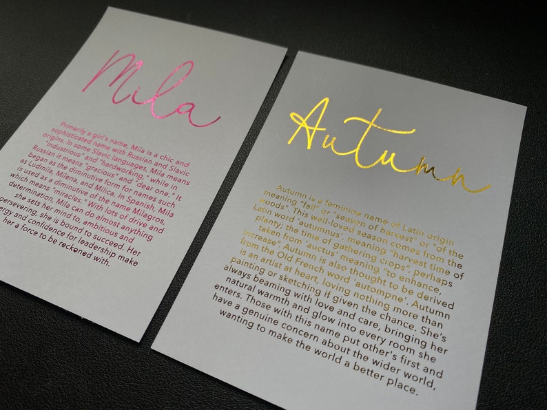 May include: Two cards with names printed in metallic foil. One card says "Mila" in pink, the other says "Autumn" in gold. Both cards have text describing the names' origins and meanings. The cards are on a gray background.