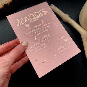 May include: A pink invitation card with gold foil text and cocktail illustrations. The card reads "MADDI'S weekend" with the date "3rd July 2025" and event details including cocktails, games, and a bottomless evening.
