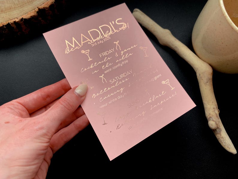 May include: A pink invitation card with gold foil text and cocktail illustrations. The card reads "MADDI'S weekend" with the date "3rd July 2025" and event details including cocktails, games, and a bottomless evening.