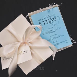 May include: A cream-colored gift box tied with a satin ribbon and a tag that says "Happy birthday." Inside, a light blue card reads "our three days in ICELAND" with a map and itinerary. The box is on a dark surface.