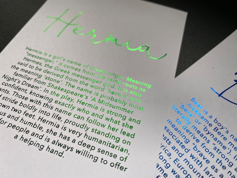May include: Two white cards with text. The card on the left has the name "Hermia" in bright green foil, with a description of the name's origin and meaning. The card on the right has the name "Bear" in blue foil, with a description of the name's origin and meaning.