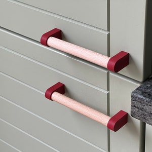 Pode incluir: Duas puxadores de armário de madeira com tampas de extremidade vermelhas. Os puxadores estão montados em uma porta de armário cinza claro.