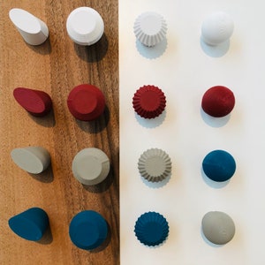 May include: An assortment of decorative knobs in various colours and shapes, including round, oval, and fluted designs. The knobs are arranged in rows on a wooden and white background.
