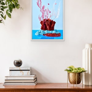 Op de afbeelding: Een ingelijste print van een stilleven met een rood koraalachtig beeldhouwwerk met roze en witte gedroogde bloemen op een blauwe achtergrond.