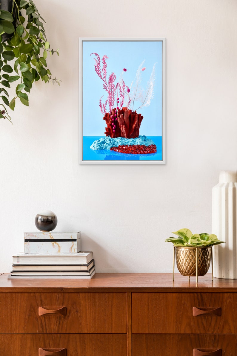Op de afbeelding: Een ingelijste print van een stilleven met een rood koraalachtig beeldhouwwerk met roze en witte gedroogde bloemen op een blauwe achtergrond.