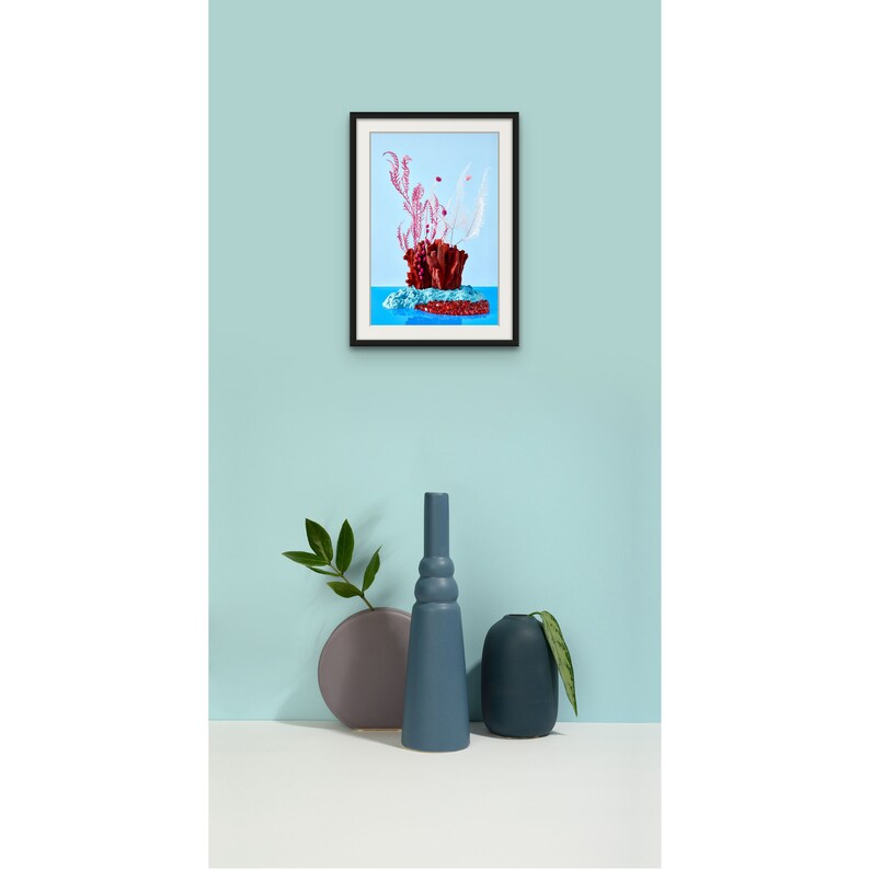 Op de afbeelding: Een ingelijste print van een rood koraalrif met roze en witte zeewier tegen een blauwe achtergrond. Twee blauwe keramische vazen en een grijze keramische vaas staan op een wit oppervlak voor de print.