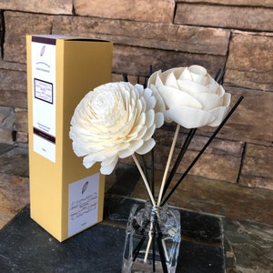 May include: A glass diffuser bottle with black reeds and two white wooden flower accents. The bottle is sitting on a dark surface and a brown box is in the background.