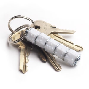 Può includere: Un mazzo di chiavi su sfondo bianco. Le chiavi sono attaccate a un portachiavi argentato. Un portachiavi bianco con superficie strutturata e fasce argentate è attaccato all'anello.